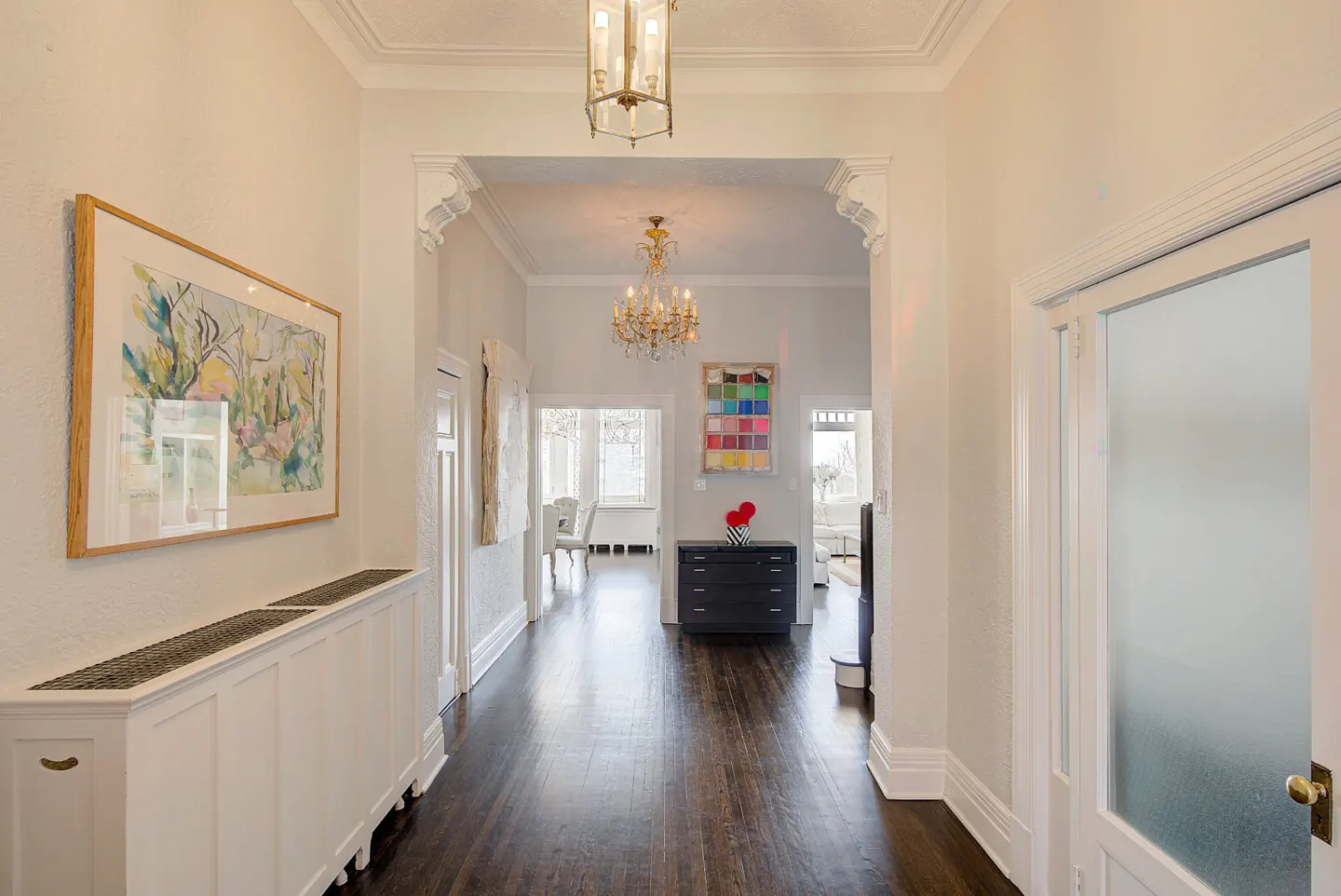 Hallway with dark wood floors, white walls, and ornate trim. A framed painting hangs above a white radiator cover. A chandelier is visible through an archway.