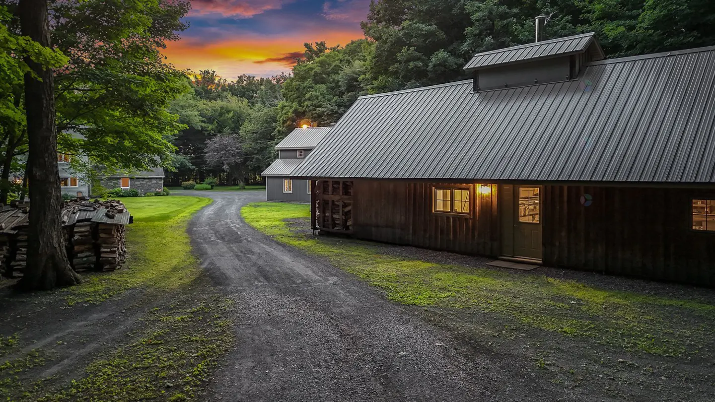 Exterior view of a rustic wooden house with a metal roof, a gravel driveway, and a sunset sky.