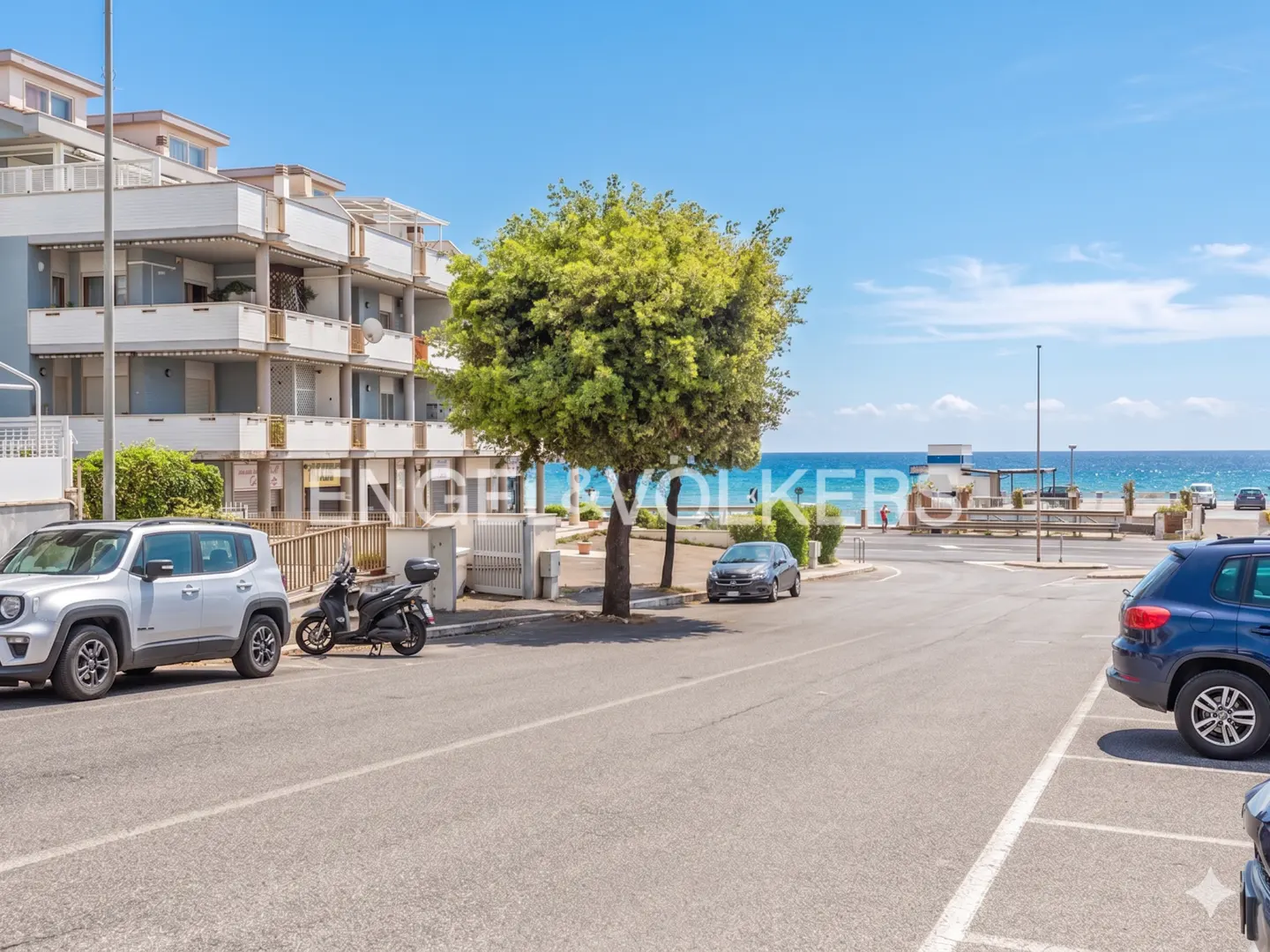 Street view of a multi-story building, cars, and a tree, with the ocean and blue sky in the background.