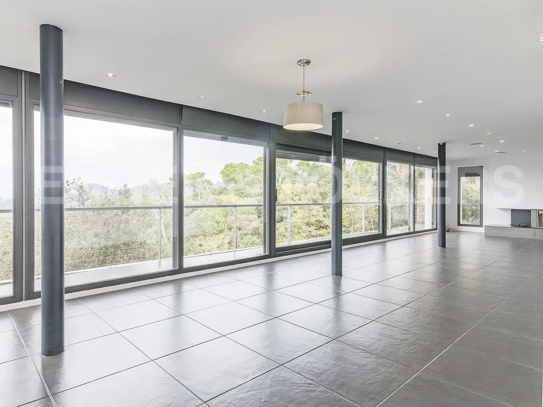 A bright, empty room with gray tile floors and floor-to-ceiling windows overlooking trees.