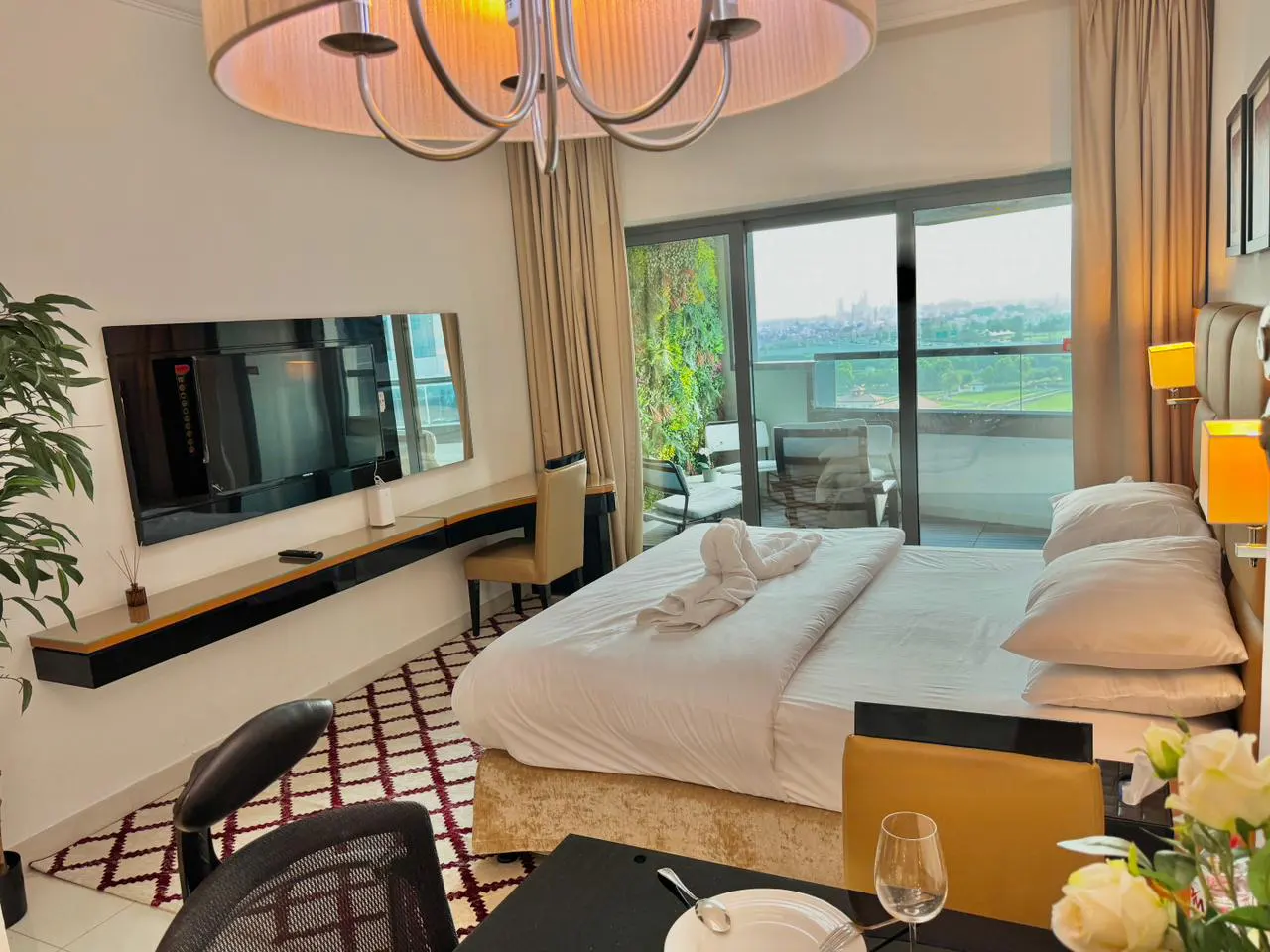 A modern bedroom with a king bed, balcony, TV, desk, and a black table with a plate and wine glass.
