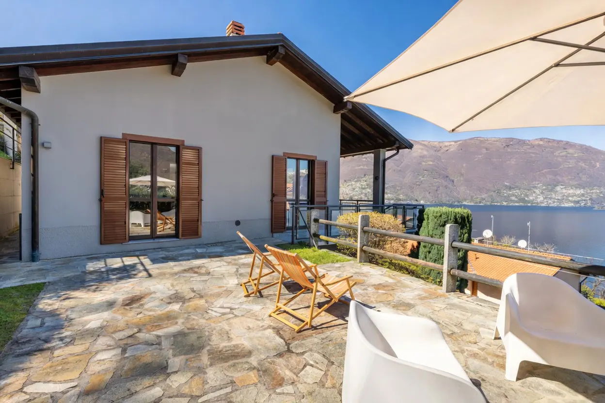 Outdoor patio with stone flooring, wooden chairs, and white loungers overlooking a lake and mountains. Cream-colored house with brown shutters in the background.