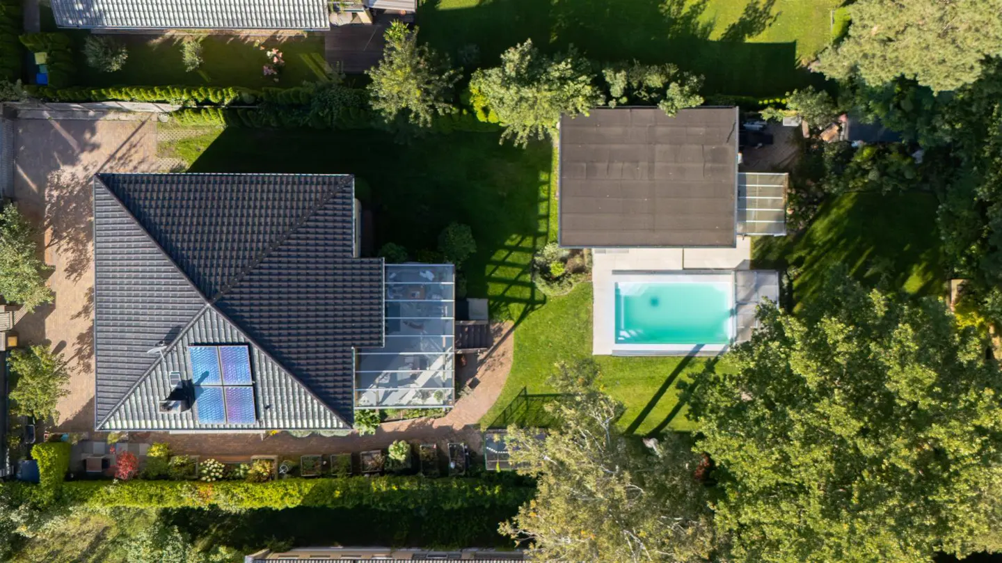 Aerial view of a house with a dark roof, solar panels, a glass-enclosed patio, and a turquoise swimming pool surrounded by green grass and trees.