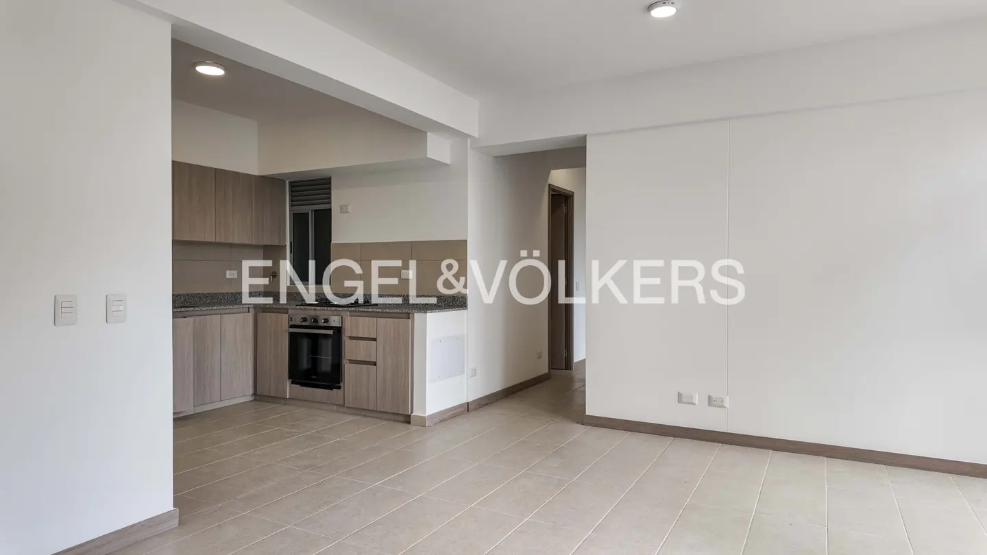 A bright, empty apartment with light wood cabinets, tile floors, and white walls. An oven is visible in the kitchen area.