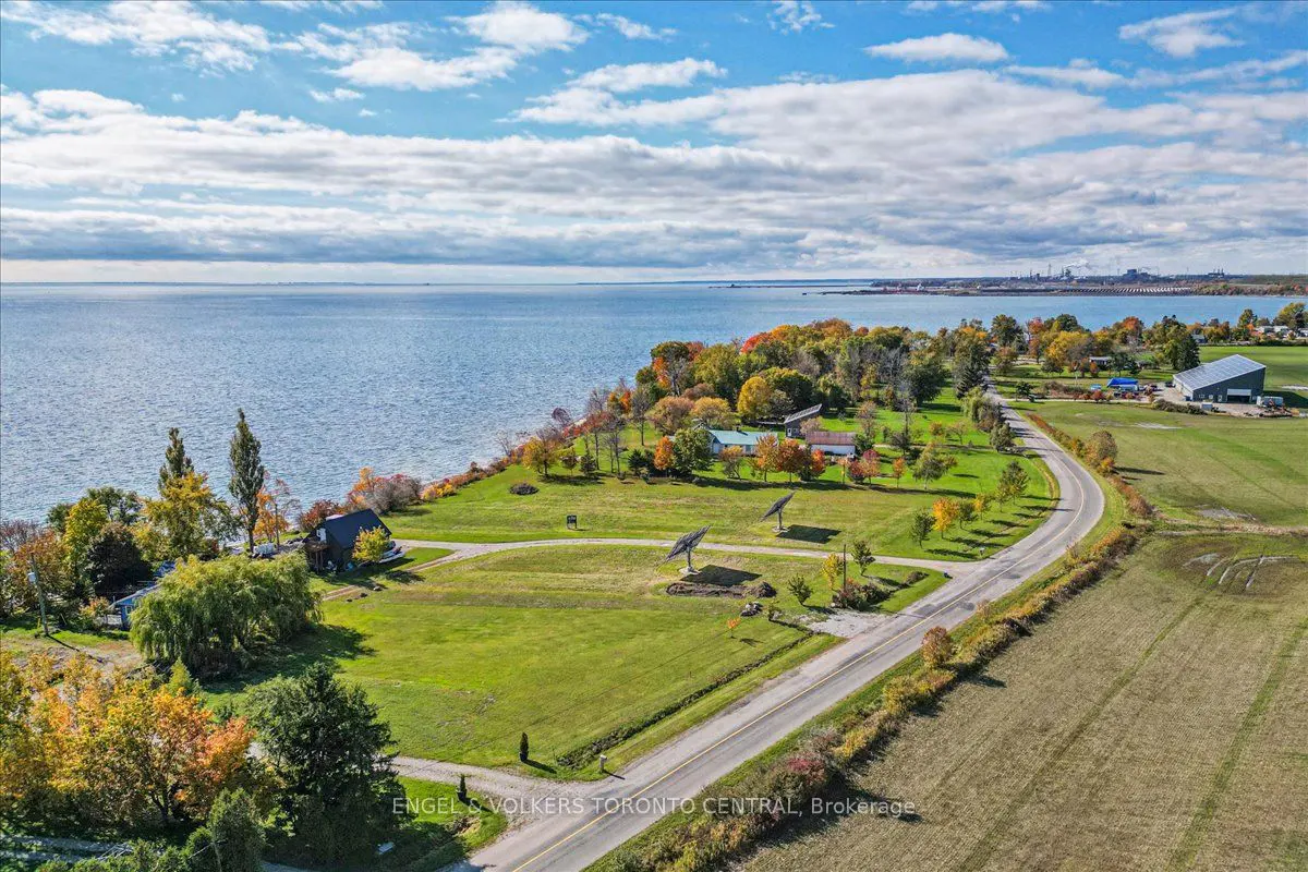 Aerial view of a waterfront property with green grass, trees with fall colors, and a road curving along the shoreline. Blue water and sky with clouds in the background.