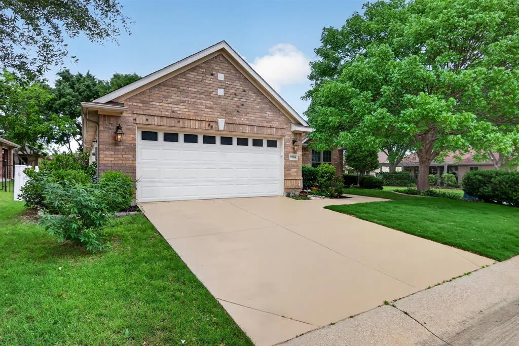 A single-story brick house with a white garage door and a concrete driveway. Green grass and trees surround the house.