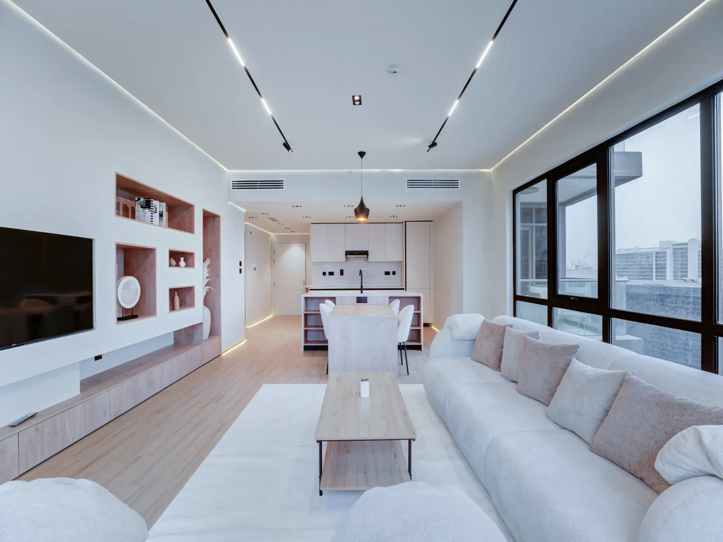 Bright, modern apartment interior with white walls, wood floors, and large windows. A white sofa faces a wood coffee table and open kitchen.