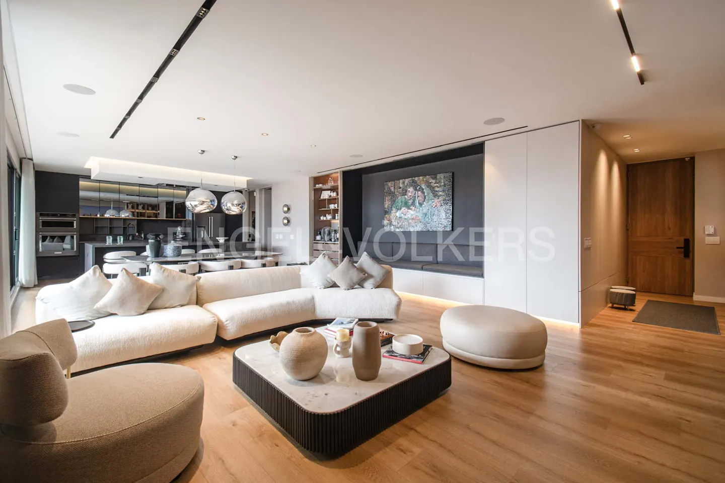 Open-concept living room with a white sectional sofa, a modern coffee table, and light wood floors. The kitchen is visible in the background.