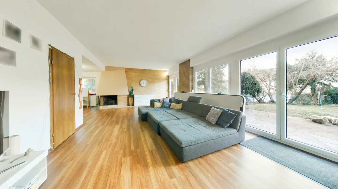 Bright living room with wood floors, a gray sectional sofa, and a fireplace. Large windows offer a view of the yard.
