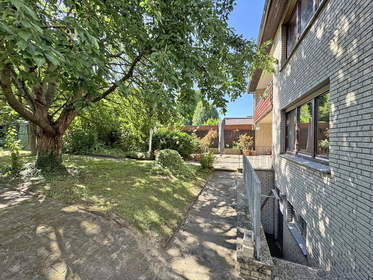 A grey brick house exterior with a side path leading to a sunny backyard featuring a large tree and a paved patio area.
