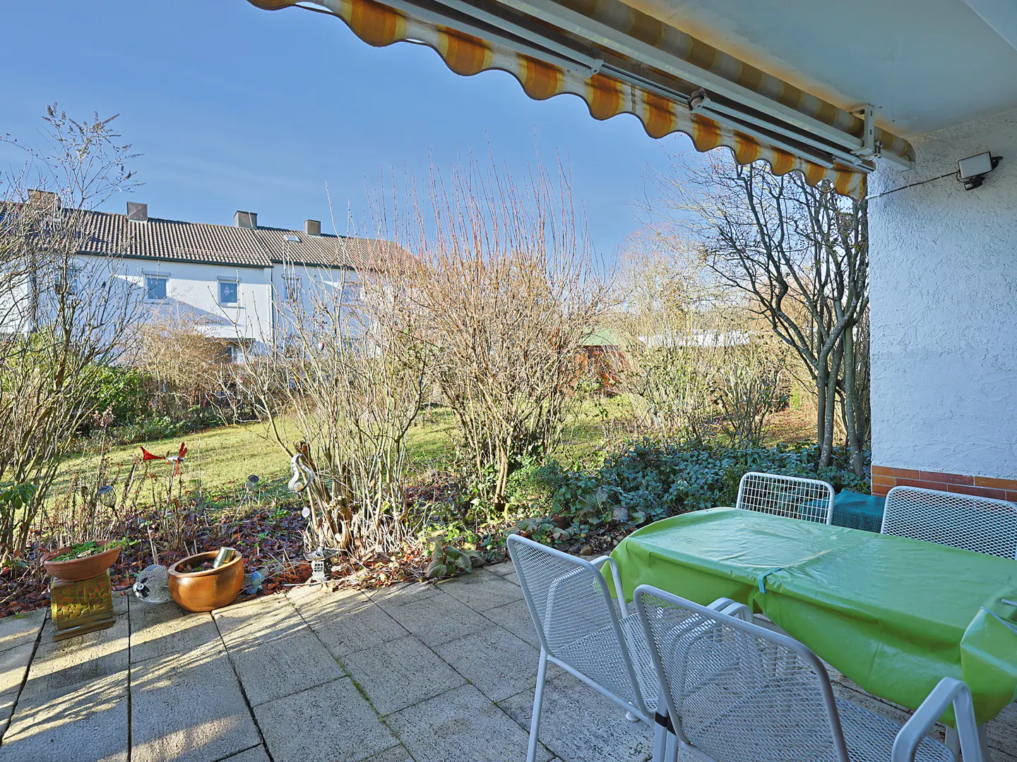 Patio view with a green-covered table and white chairs. A garden with trees and a house are in the background.