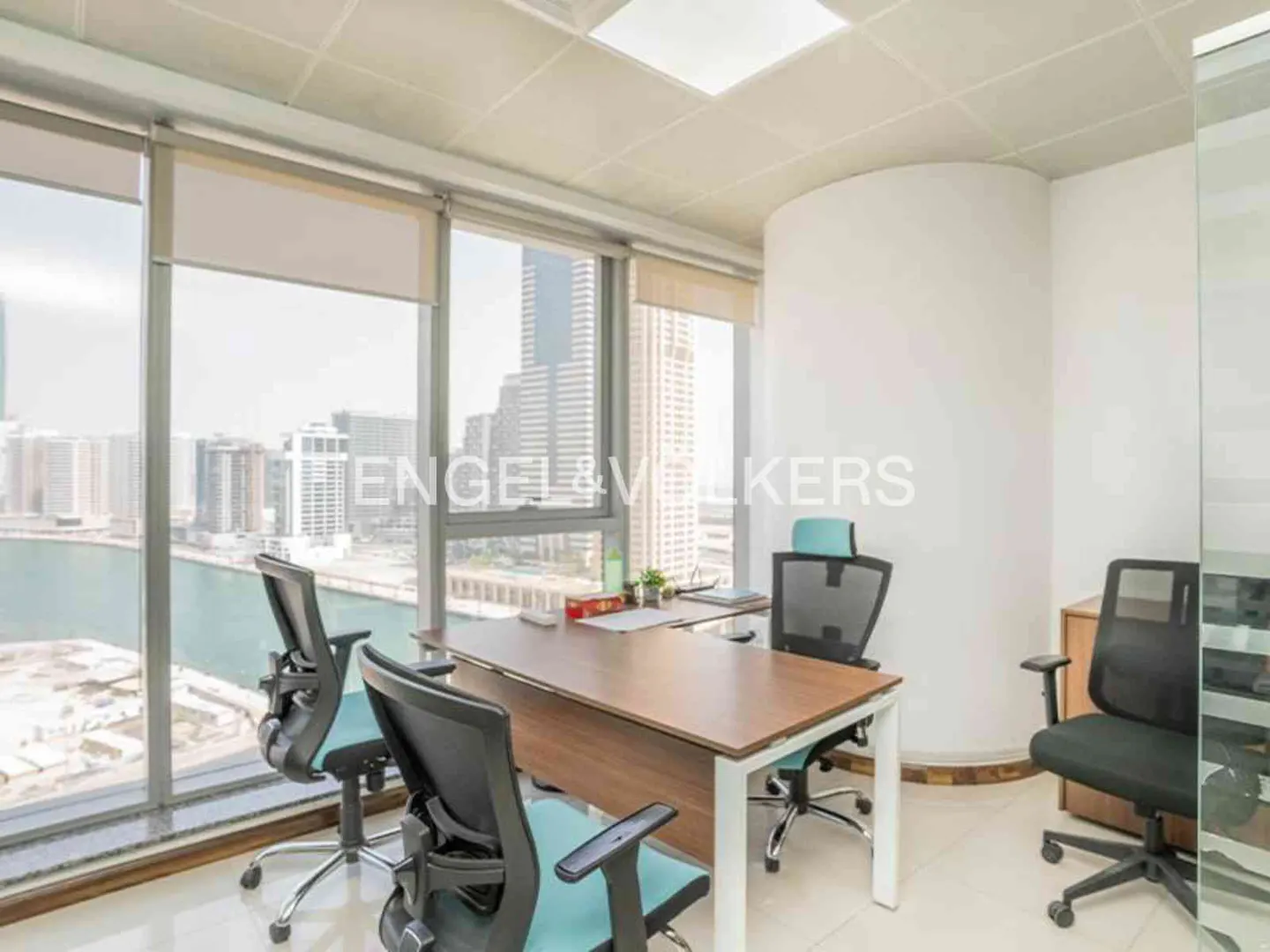 Bright office space with a large window overlooking a city skyline and water. A wooden desk with chairs faces the view.