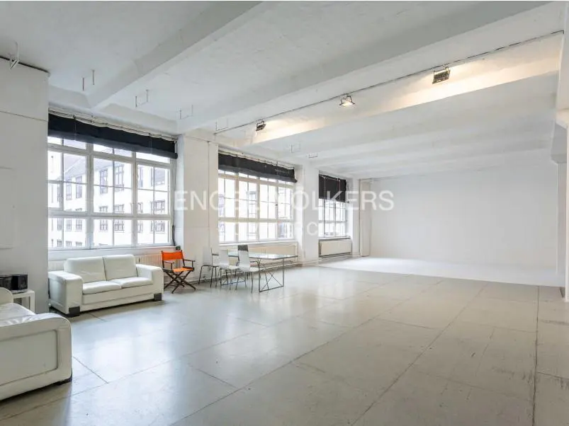 Bright, spacious studio apartment with white walls, concrete floors, and large windows. Sofas and chairs are arranged near the windows.