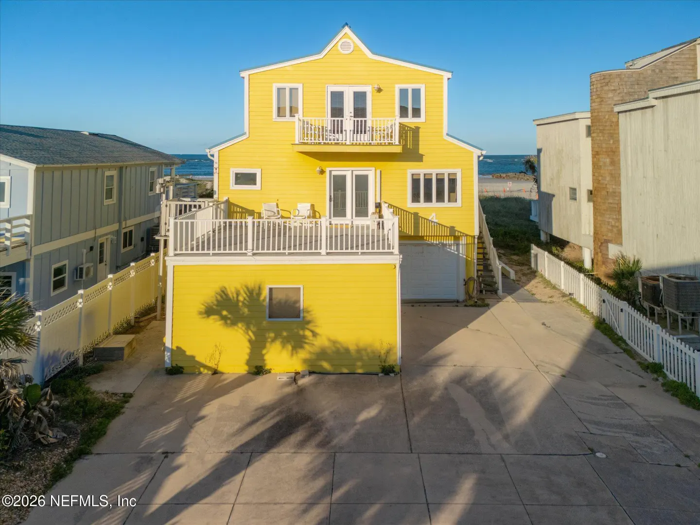 Bright yellow three-story beach house with a large deck, concrete driveway, and ocean views in the background.