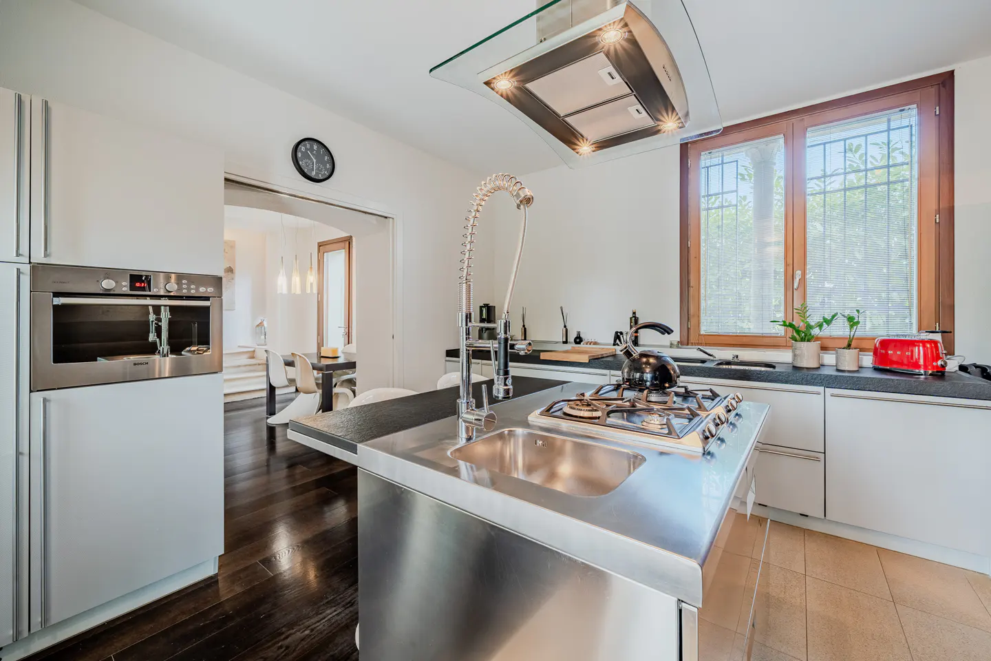 Bright kitchen with stainless steel island, sink, and gas stove. White cabinets, dark countertops, and a window with a wooden frame.