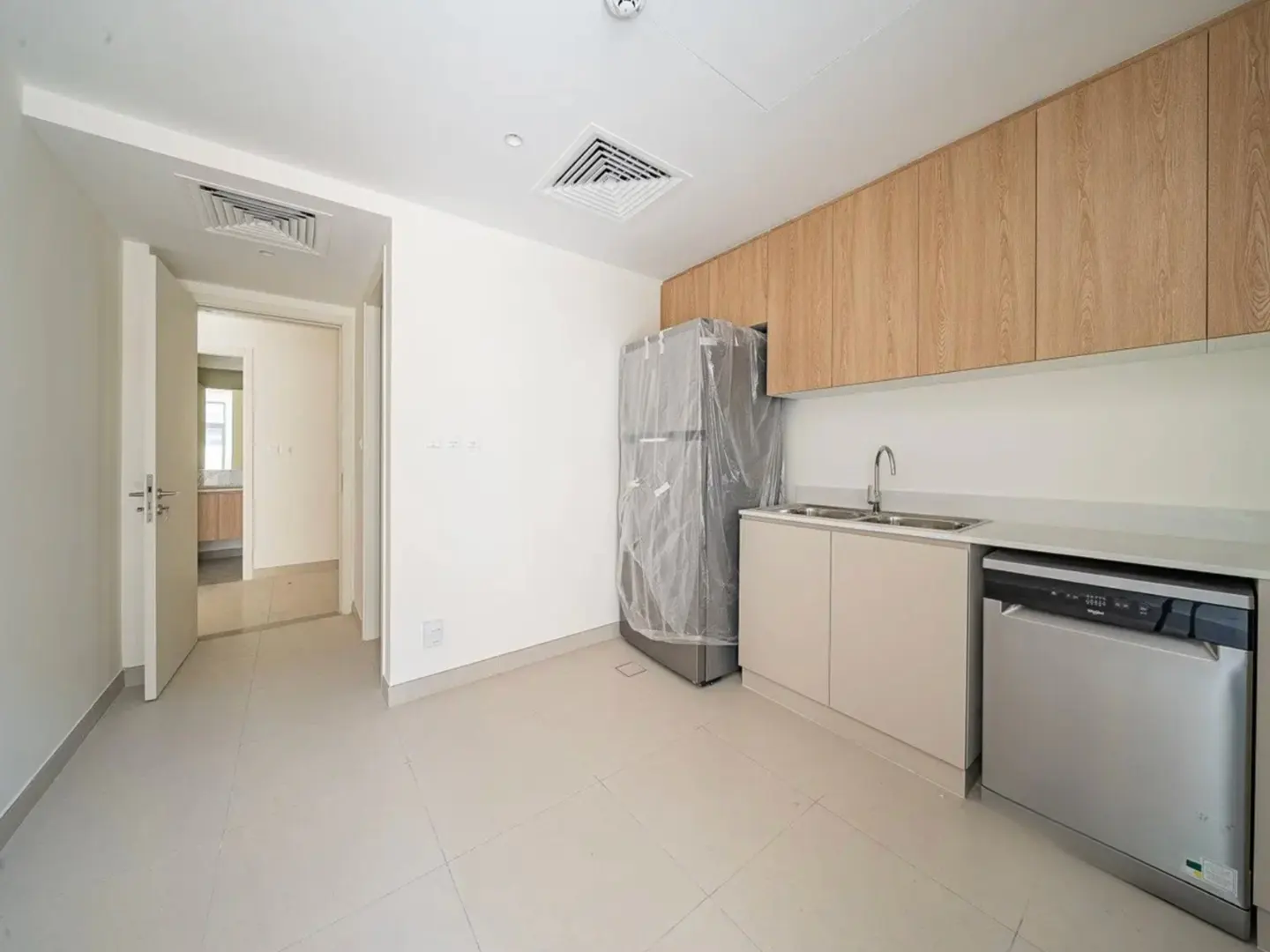Bright, empty kitchen with light wood cabinets, stainless steel appliances, and a refrigerator wrapped in plastic. A doorway leads to another room.
