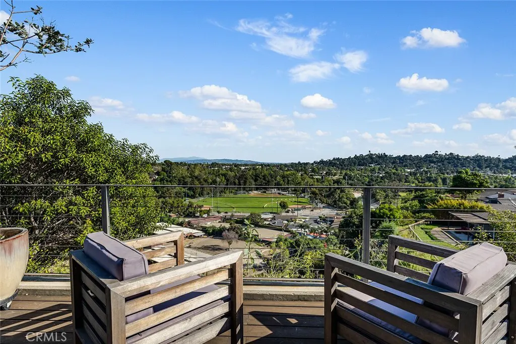 Wooden chairs on a deck overlook a green field and cityscape under a blue sky with scattered clouds.