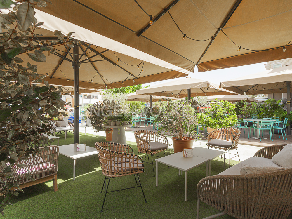 Outdoor patio with tan umbrellas, wicker chairs, and green turf. Tables are white, and plants add greenery to the space.