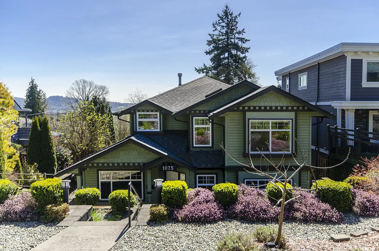Two-story green house with dark trim and a gray roof. The house number "1075" is visible above the front door. The yard has purple flowers and green bushes.