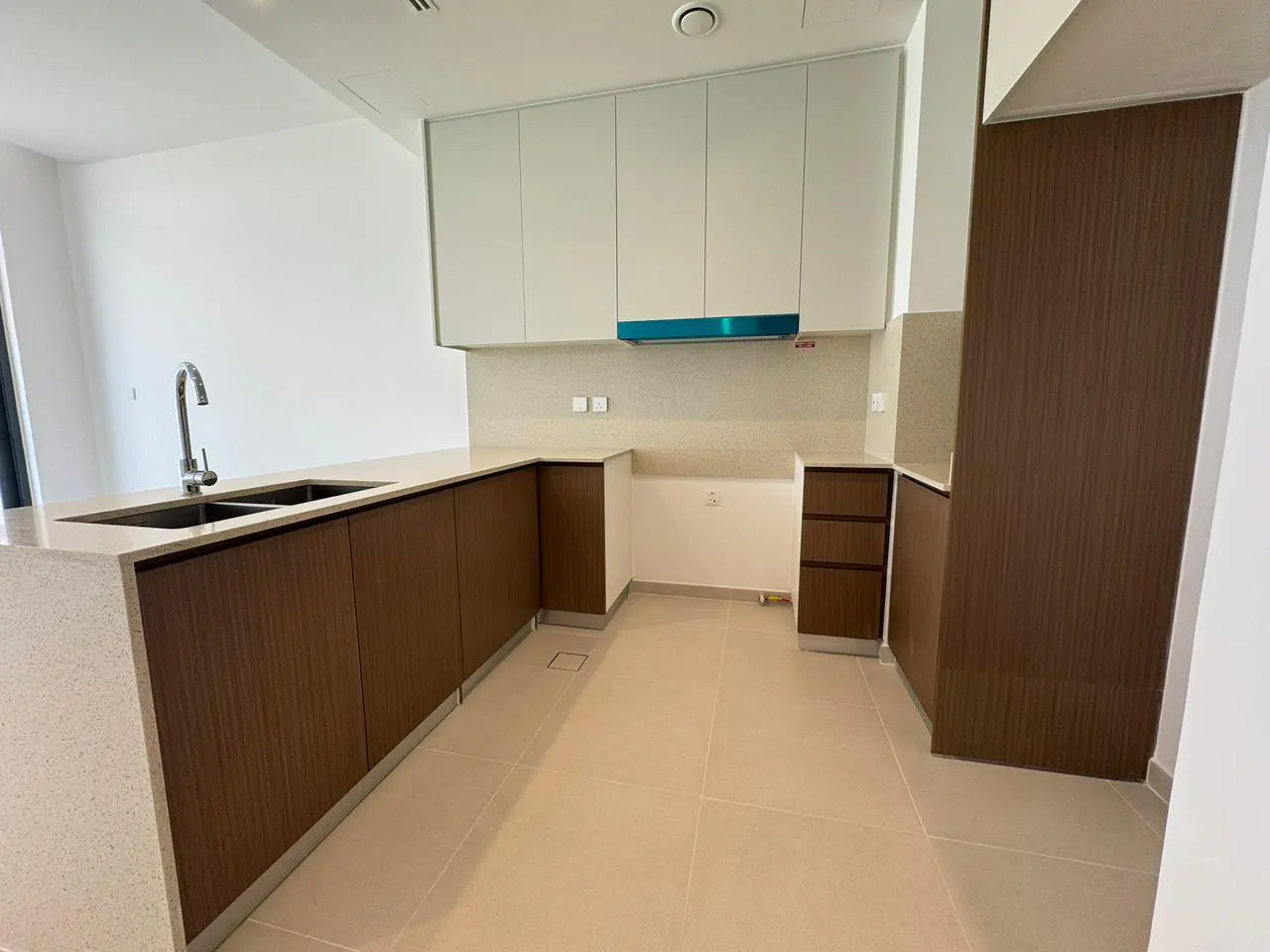 A modern kitchen with white upper cabinets, wood-grain lower cabinets, and a speckled countertop. A stainless steel sink and faucet are visible.