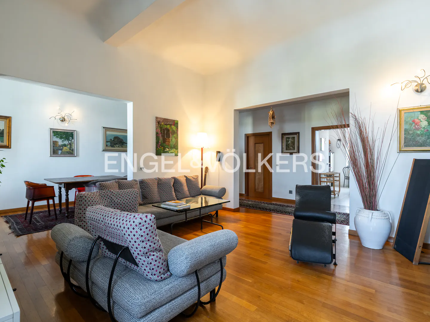 Bright living room with hardwood floors, gray sofas, glass coffee table, and black leather chair. Artwork adorns the white walls.