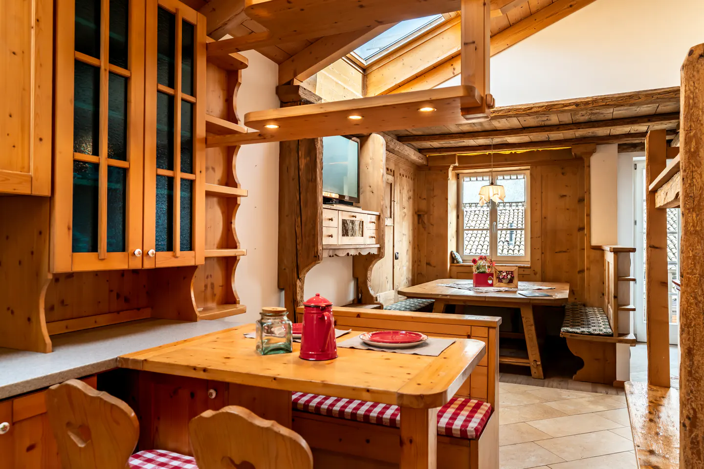 Rustic kitchen with light wood cabinets, a dining table with red-checkered cushions, and a bright, cozy atmosphere.