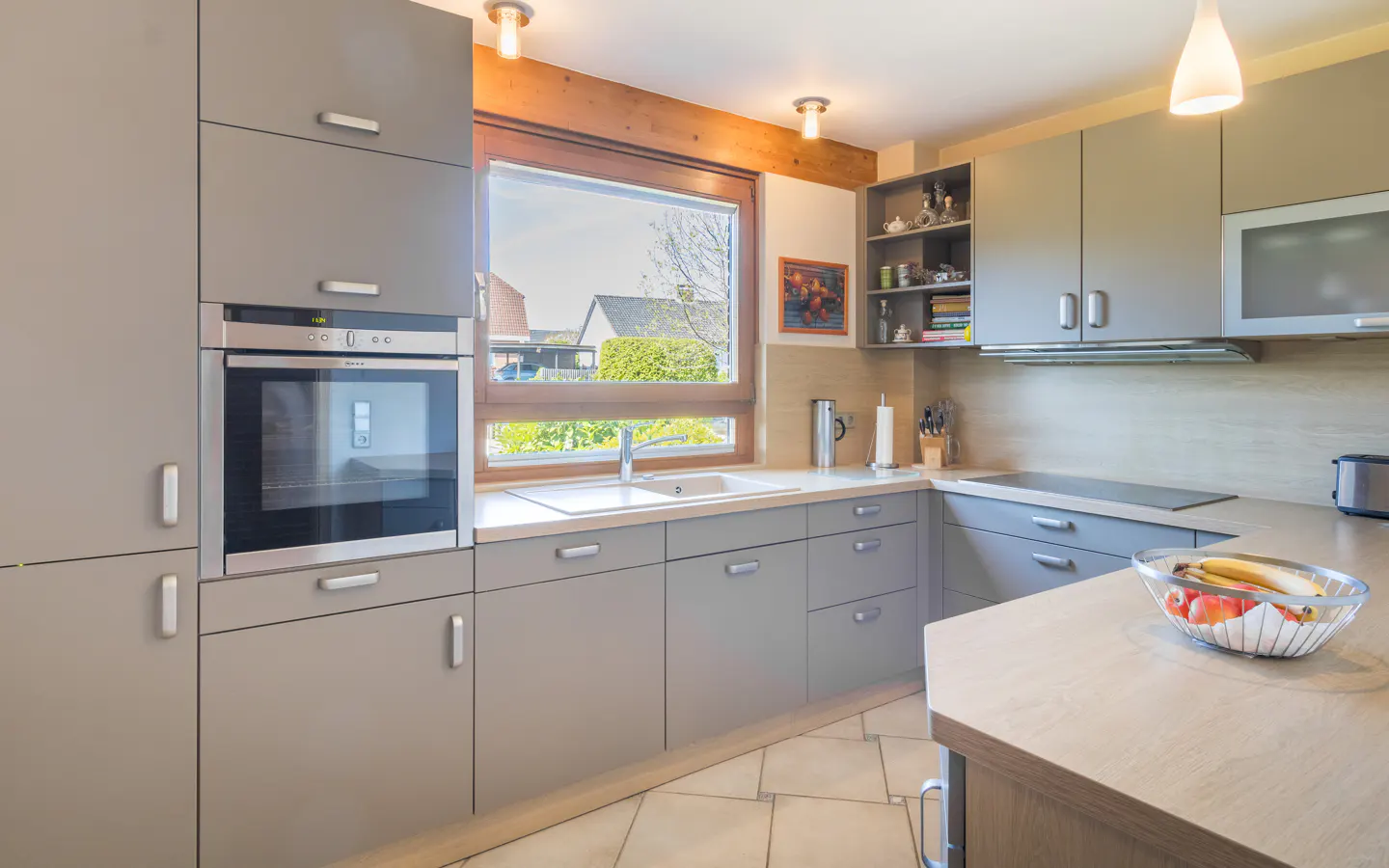 A modern kitchen with gray cabinets, stainless steel appliances, and a window overlooking a green yard. A bowl of fruit sits on the countertop.