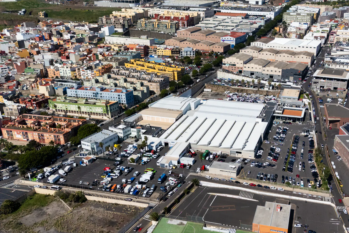 Aerial view of a large, white-roofed industrial building with parking lots, surrounded by colorful residential and commercial buildings.