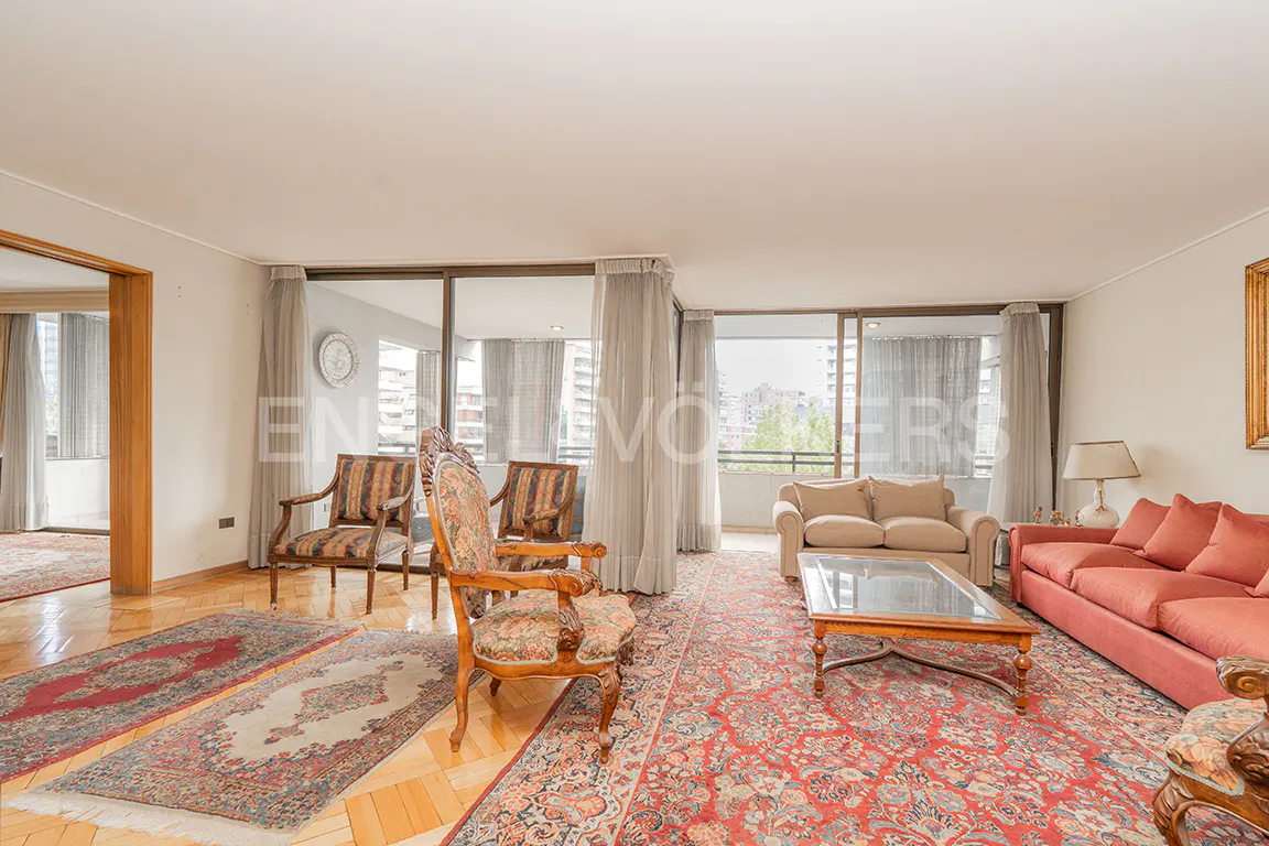 A bright living room with hardwood floors, patterned rugs, and floor-to-ceiling windows. Furniture includes a red sofa, beige loveseat, and wooden chairs.
