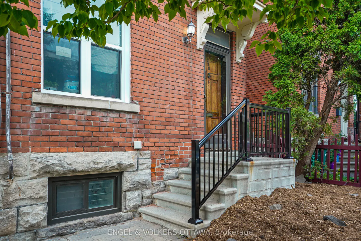 Brick house exterior with stone foundation, steps leading to a wooden door with black railing. Green tree on the right.