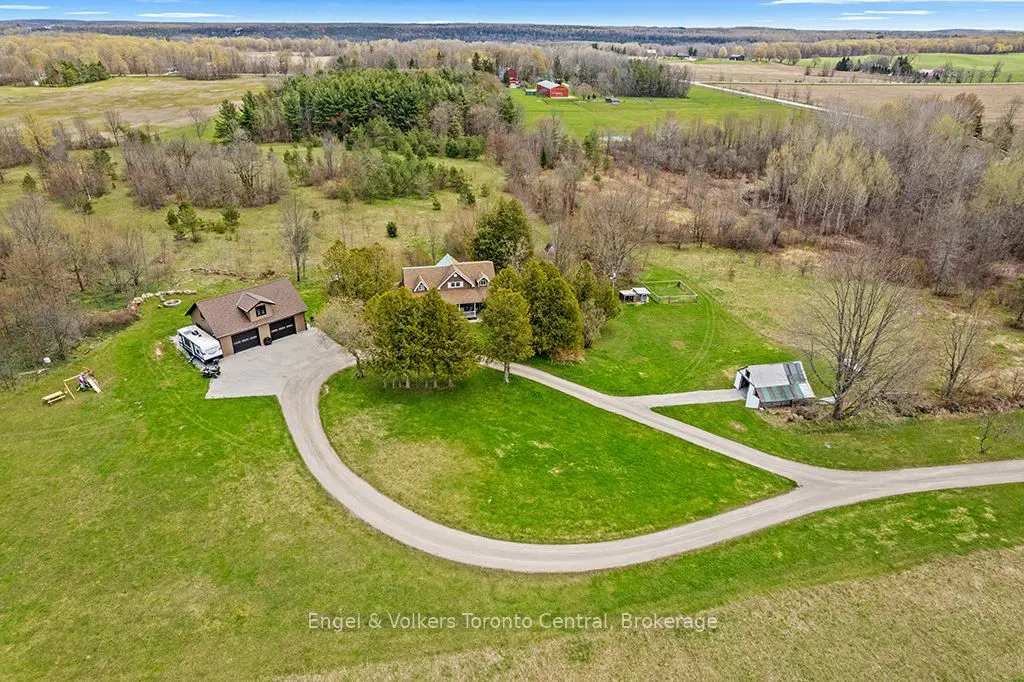 Aerial view of a rural property with a house, garage, and barn surrounded by green lawns and trees. A winding driveway leads to the buildings. Fields and forests are in the background.
