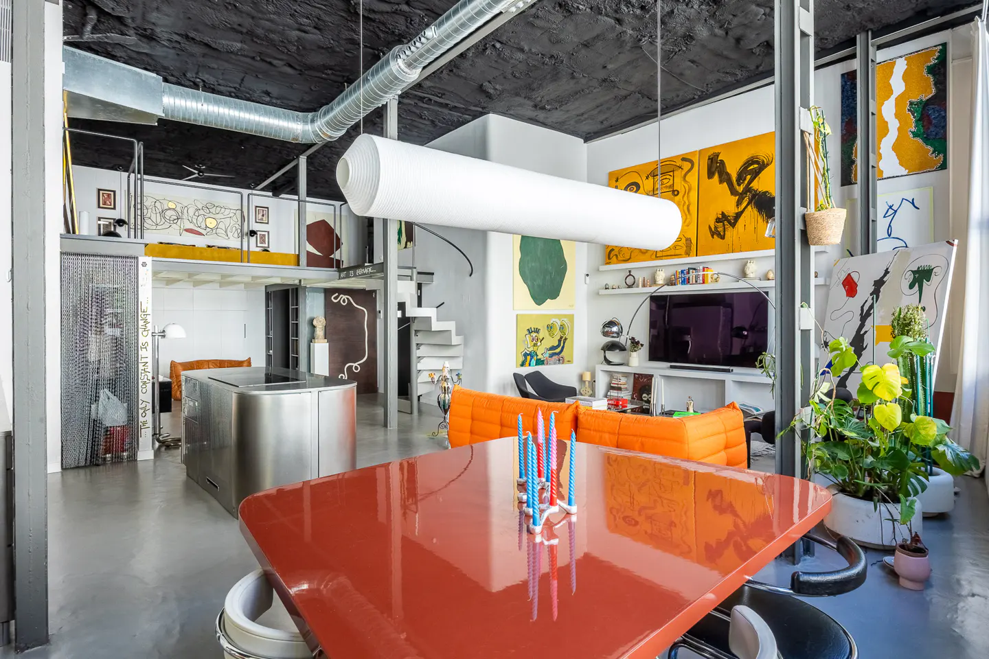 A modern loft apartment with a red table, orange sofa, and abstract art. A white cylindrical light hangs from the black ceiling.