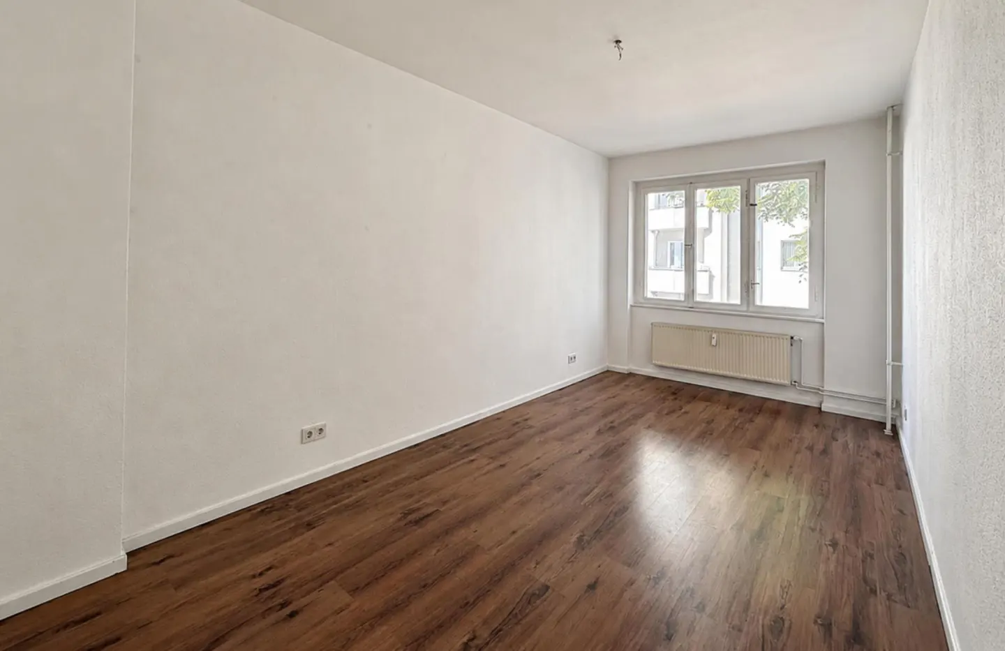 Empty room with white walls, dark wood-look flooring, a window, and a radiator under the window.