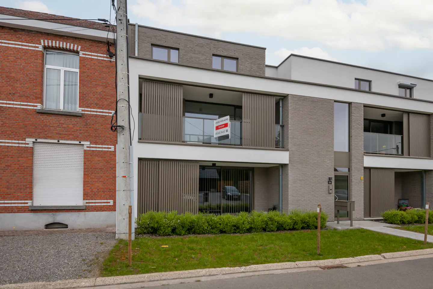 A modern, two-story apartment building with a "For Sale" sign on the balcony. The building is gray brick with white trim and brown vertical blinds. A red brick building is on the left.