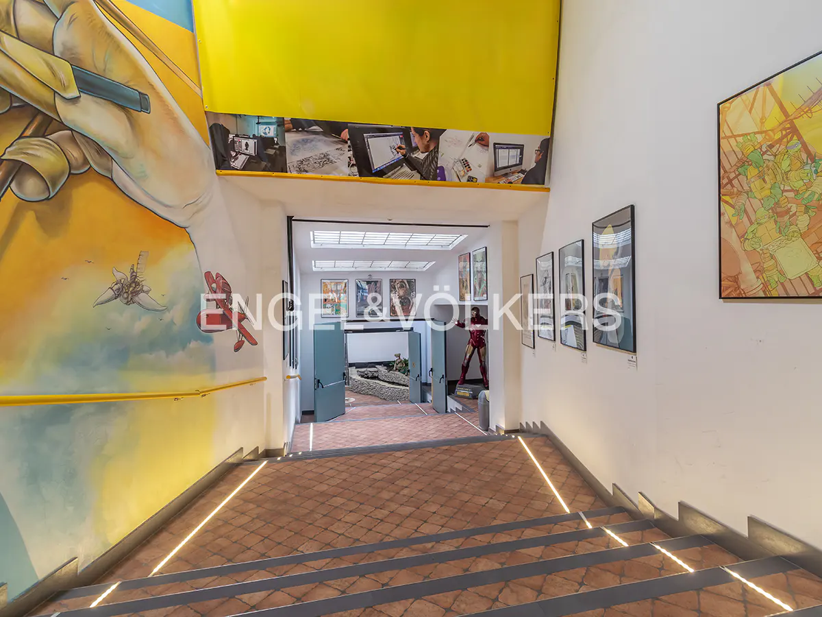 Stairway leading to a comic-themed room with art, figures, and a mural of a hand drawing. The walls are white with yellow accents.