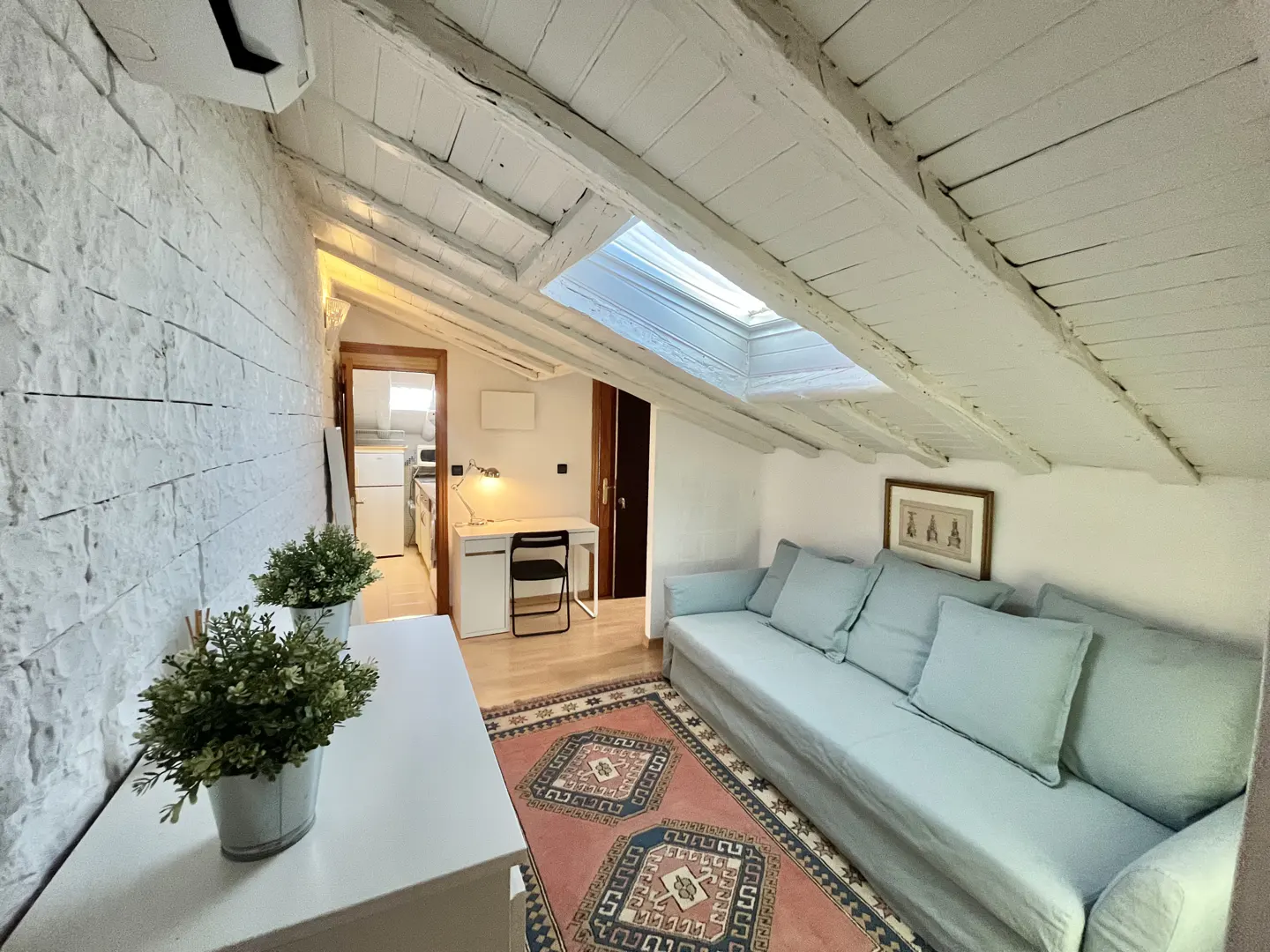 Attic living room with white brick wall, light blue sofa, patterned rug, and a desk area under a sloped wooden ceiling.