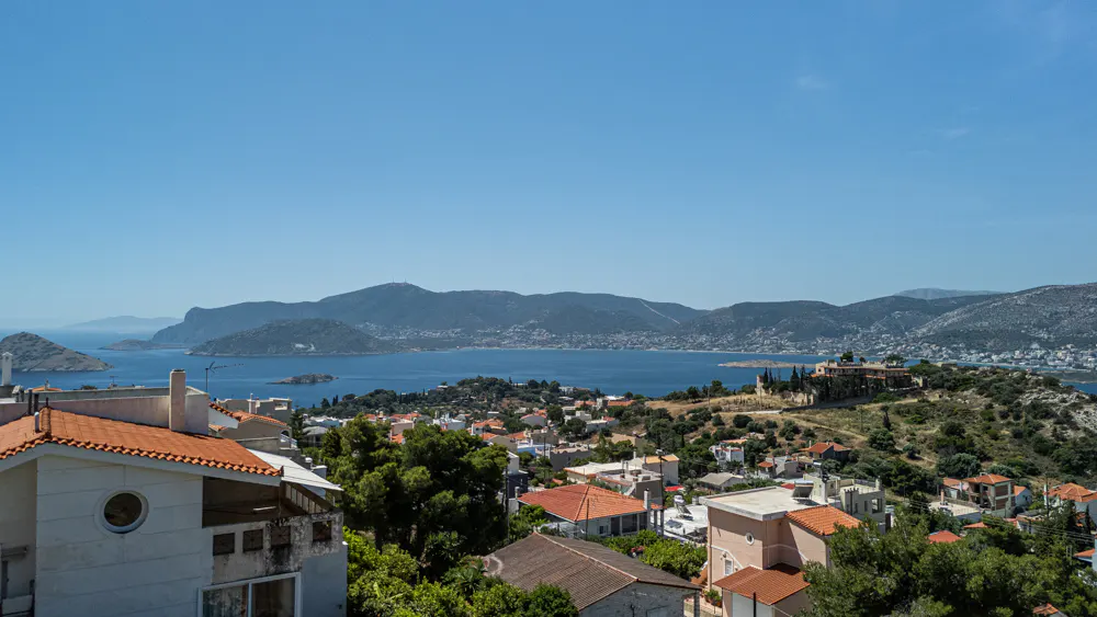 Elevated view of a coastal town with terracotta-roofed houses, lush greenery, and a blue bay surrounded by mountains.