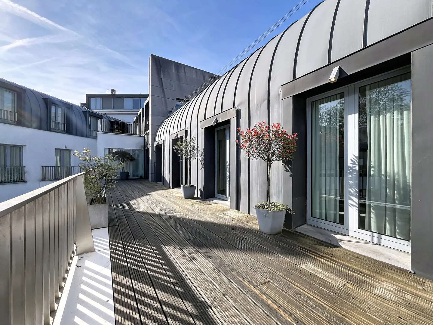Exterior view of a modern building with a wooden deck, potted plants, and gray metal siding under a blue sky.
