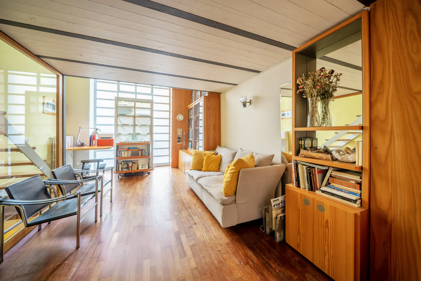Bright living room with wood floors, a beige sofa with yellow pillows, and built-in wood shelving. Two black chairs sit near a glass wall.