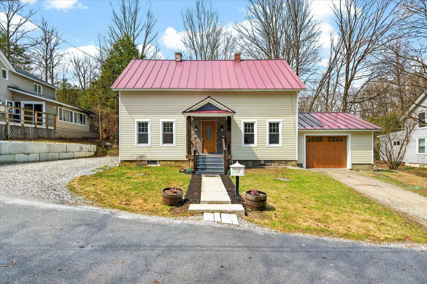 Beige house with a red roof and a detached garage on a green lawn with trees in the background.