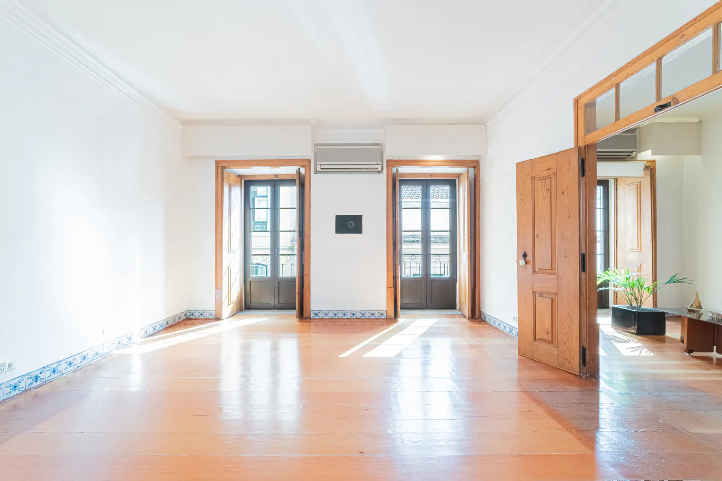 Bright, empty room with wood floors, white walls, and two sets of open, dark-framed doors leading to balconies. Ajar wood door on the right.