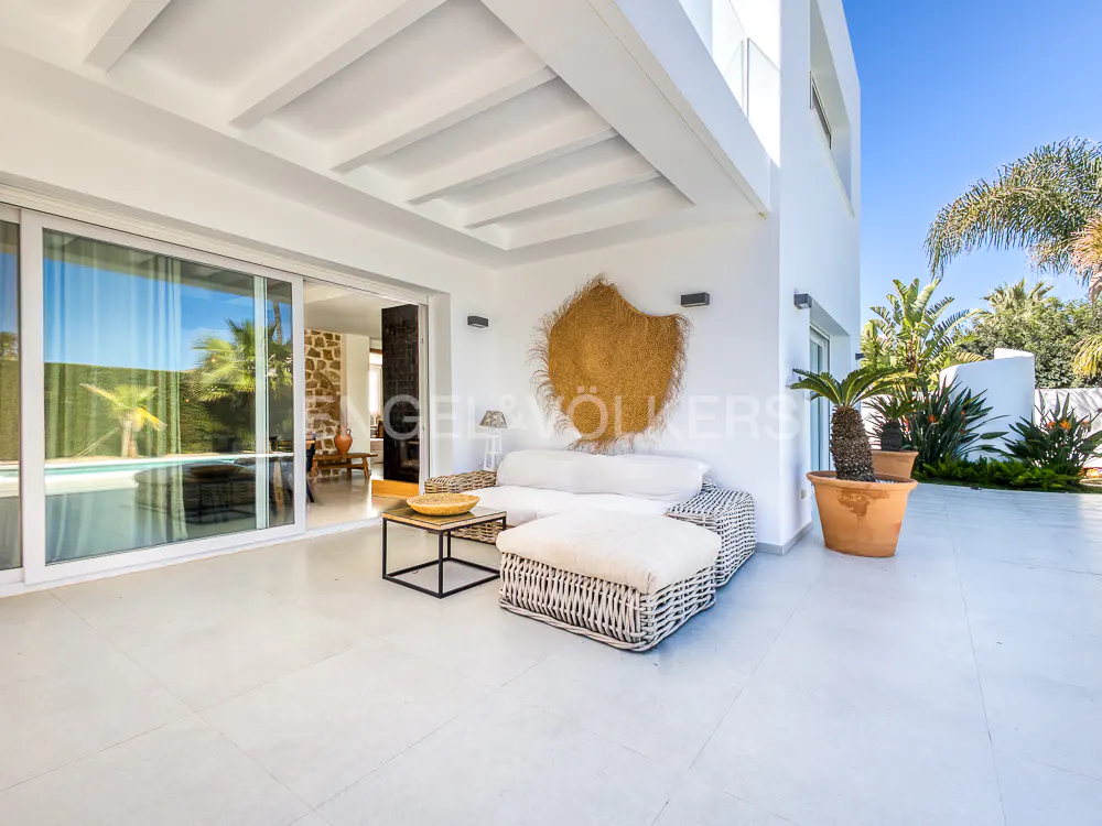 Outdoor patio with white wicker furniture, a straw wall hanging, and a potted palm tree. Sliding glass doors lead to the interior.
