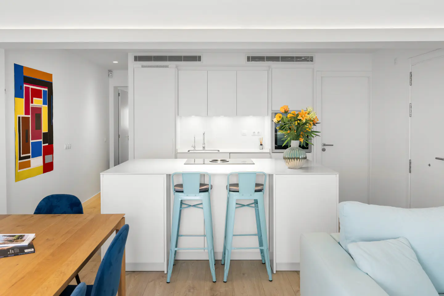 Bright, modern kitchen with white cabinets, island, and blue stools. A colorful abstract painting hangs on the wall.