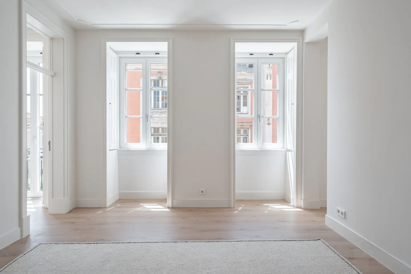 Bright, minimalist room with white walls, light wood floors, a neutral rug, and two windows overlooking a city building.