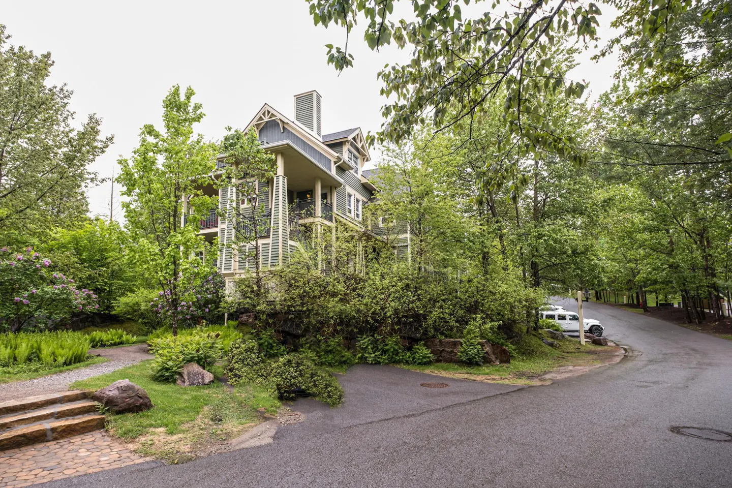 A two-story green and gray house is surrounded by lush green trees and bushes. A paved road curves in front of the house.