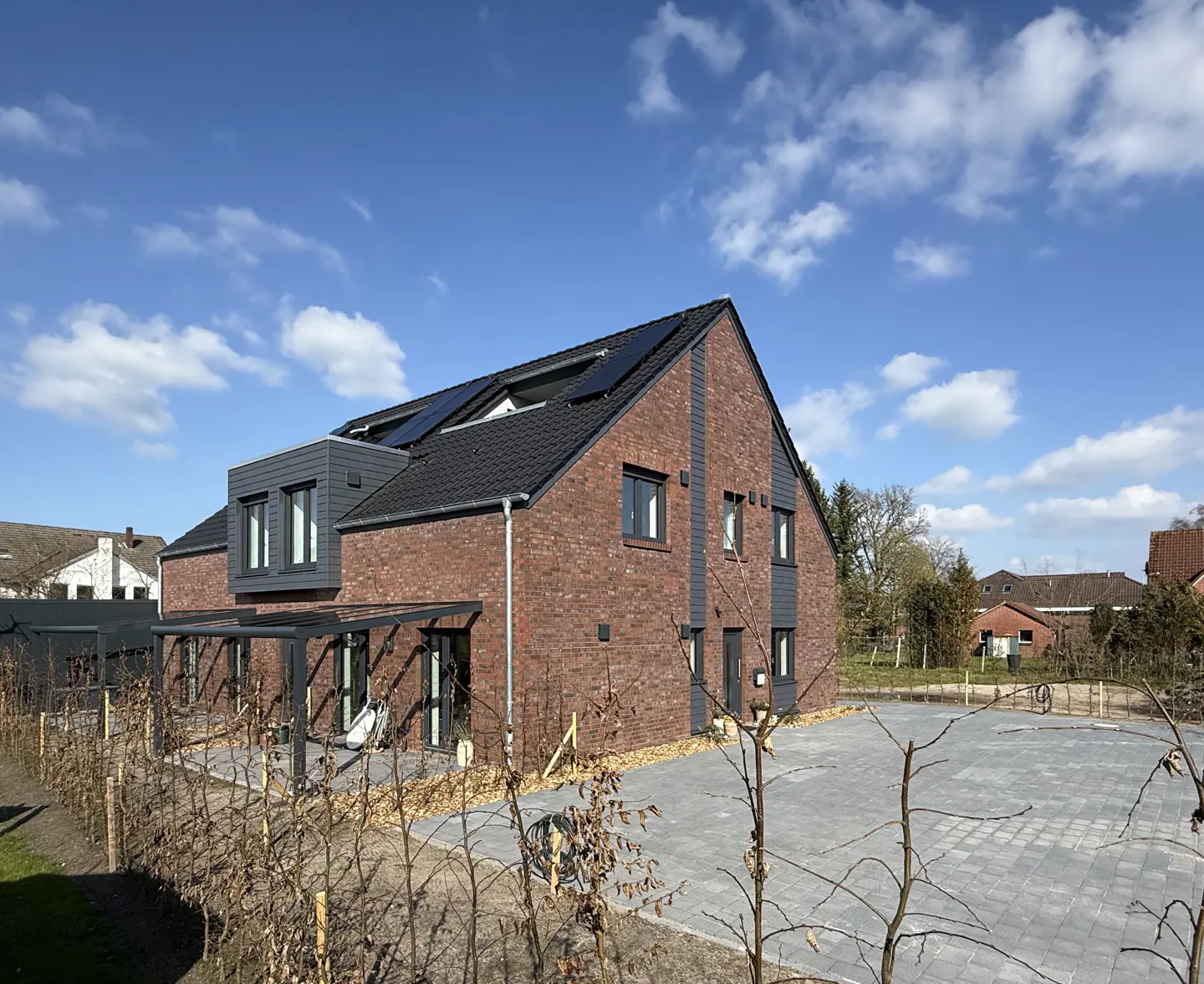 A modern two-story brick house with a black roof and solar panels under a blue sky. A gray carport is attached to the side.