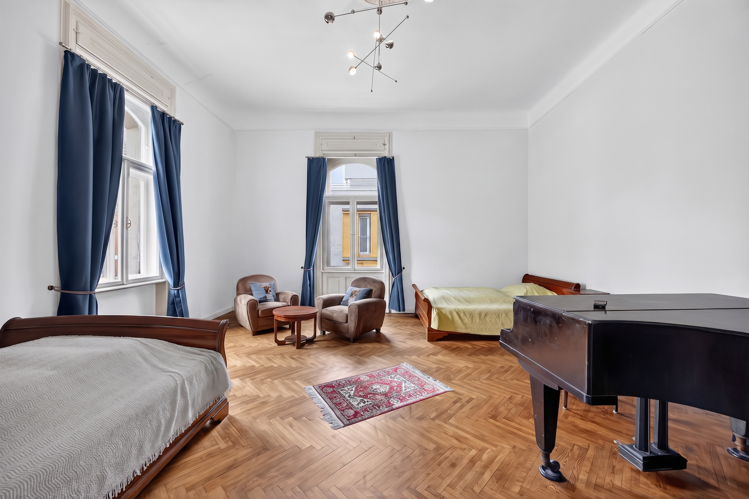 Bright room with herringbone wood floors, two beds, a black piano, and two brown armchairs with a small table. Blue curtains hang by the windows.