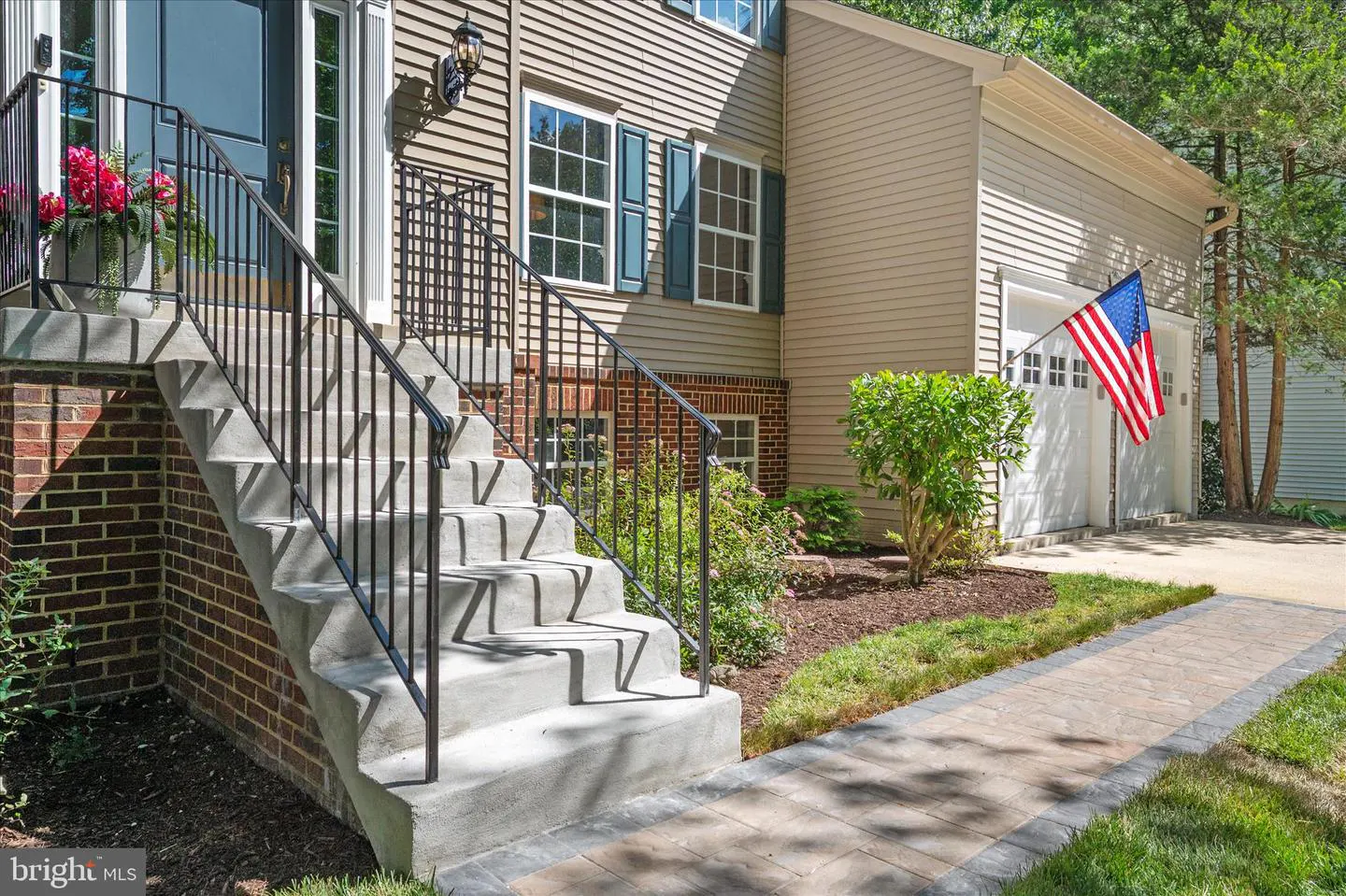 Concrete steps with black railings lead to a house with tan siding, blue shutters, and an American flag by the garage.