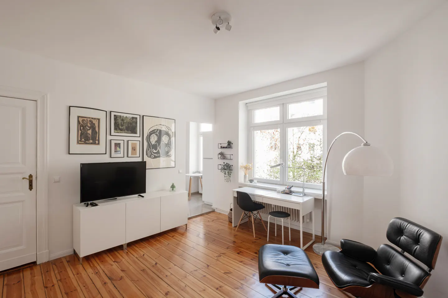 Bright living room with wood floors, white walls, and a window. A desk, chair, and black leather chair with ottoman are present. Artwork hangs above a white media console.