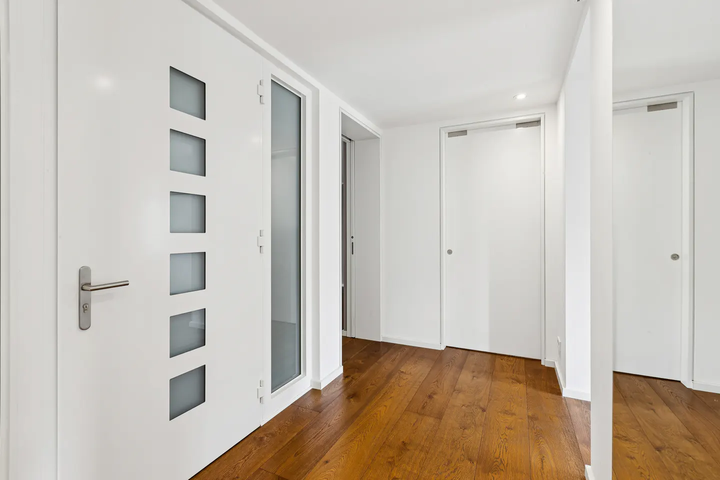 Hallway with white walls, brown wood floors, and white doors. One door has frosted square windows.